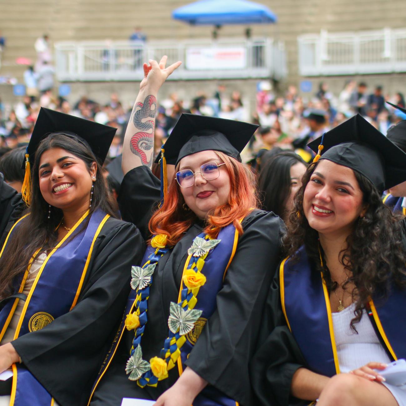 Three female students in graduation robes and caps smile and hold up peace fingers